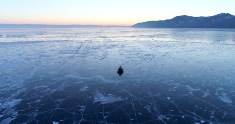 Aerial: Hovercraft travels on frozen Lake Baikal by mountains against orange sky