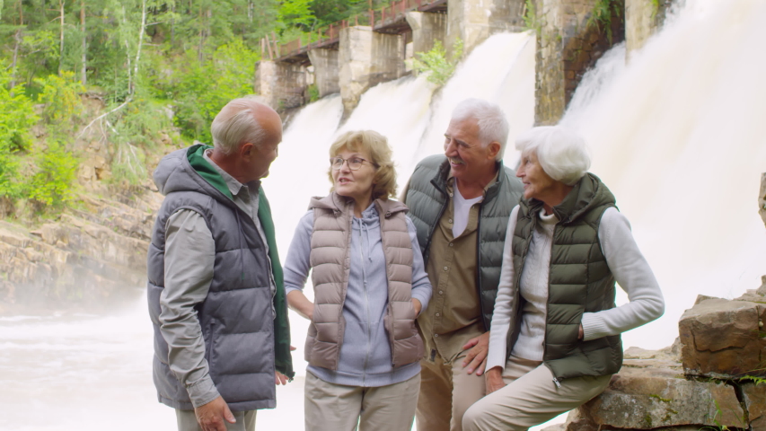 Handheld medium shot of happy senior people standing in front of dam spillway and chatting, then looking at camera and smiling