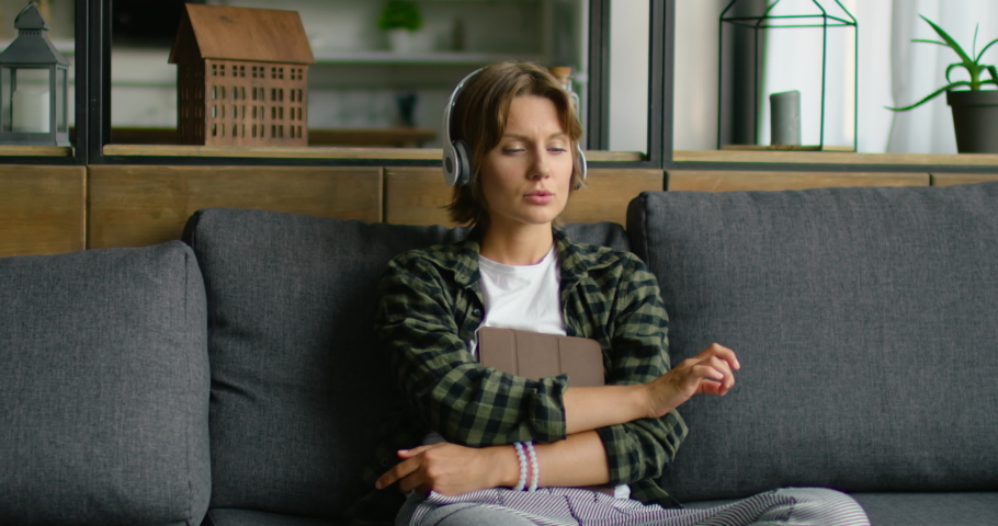 Young woman is listening soft music in wireless headset, holding tablet computer, gadget, relaxing on sofa, enjoying life style, in romantic mood, indoor. Caucasian, Middle plan, 4K, RED camera.
