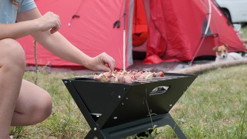 Woman's hand overturns a kebab barbecue on small brazier against red tent, dog Jack Russel Terrier in clearing with green grass. Family summer vacation camping with tent. Lifestyle