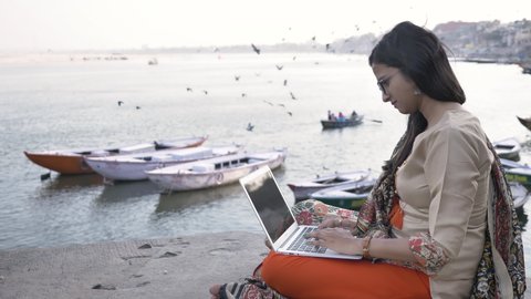 A young and beautiful woman dressed in traditional salwar kameez sitting on a platform working on a laptop next to a river, while boats are docked and birds are flying in the background during sunset.