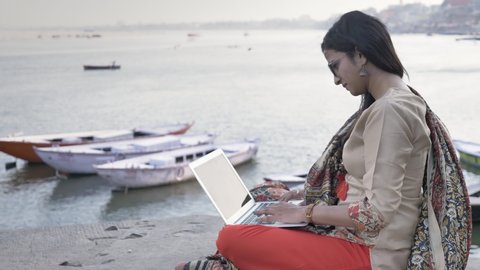 A movement shot of a young and beautiful woman dressed in traditional salwar kameez sitting on a platform working on a laptop next to river, boats are seen in the background during sunset.