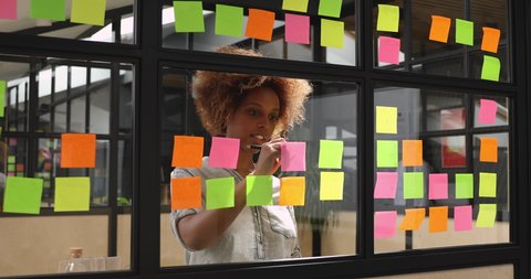 Focused young african female professional student worker coach write ideas replacing post it sticky notes with done research tasks planning project strategy on glass scrum board wall in modern office