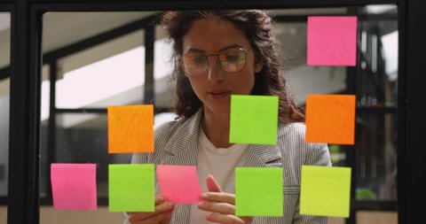 Focused young business woman think of project strategy attach replace sticky post it notes on glass board, female corporate leader coach planning organize work on stickers on scrum glass window wall