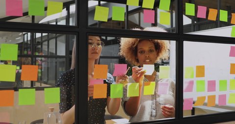 Two african and asian businesswomen talk work together in teamwork share creative ideas discuss project plan replace post it sticky note stand behind glass discuss corporate strategy at team briefing