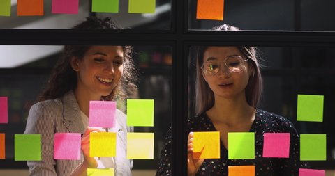 Happy friendly diverse female colleagues talking working with sticky notes, smiling professional team two businesswomen sharing creative ideas replacing stickers planning project write on glass wall