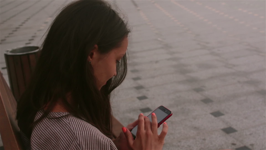 Young beautiful girl sitting on a bench in the park, smiling and writing a message on the internet chatting on her smartphone.