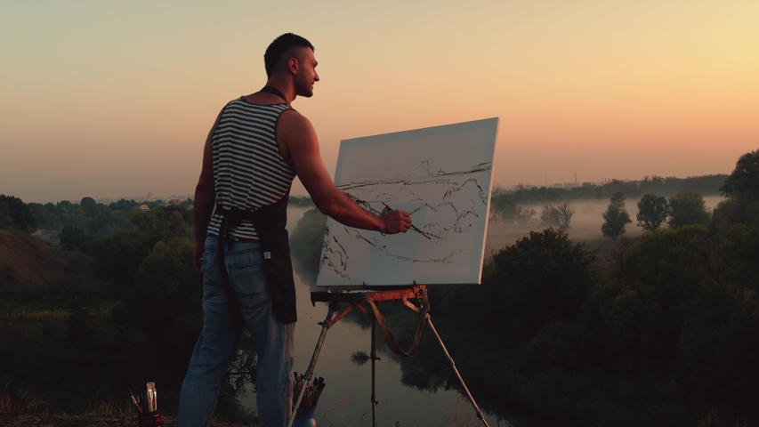 Landscape painter. Young man painting a natural scenery with oil paints. Morning sunrise or sunset