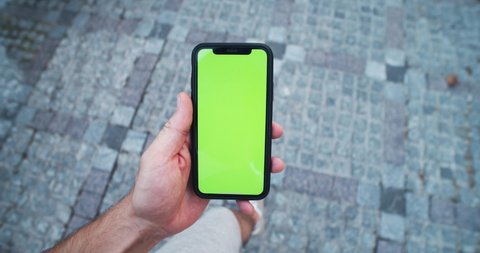 Kyiv, Ukraine - August 20, 2019: Young caucasian man using smartphone walking in the street close-up. First-person view of male hand swiping on mock-up mobile greenscreen device outdoor.