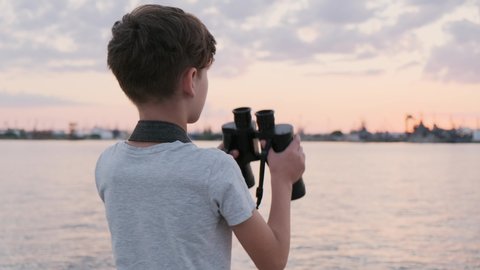 Happy boy holds binoculars stands and looks into distance at bright sunset and shows with his hand at sun on seaport pier. Quiet expanse of water. Children's summer vacation. Travel. Tourism