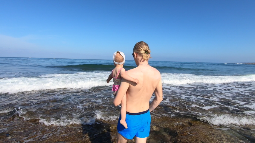 Young father hold his little daughter on the hand and show beautiful sea with big wave. Active family travel to Cyprus, Paphos and spending time together