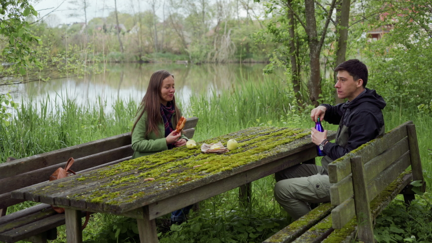 Girl and guy have picknic with apples, benzel and hot tea in thermos. They sitting on old wooden benchs and table which are covered with green moss