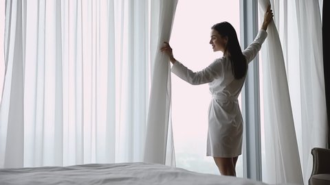 Happy confident rich young woman wear nightgown come to big window open curtain lace at modern home hotel enjoy good morning city view dream or future start day after wake up in bedroom, slow motion