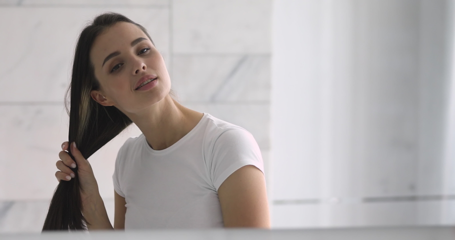 Smiling attractive young woman brushing hair looking in the mirror, happy confident young beautiful woman doing hairstyle in bathroom bedroom in morning get ready during everyday routine at home