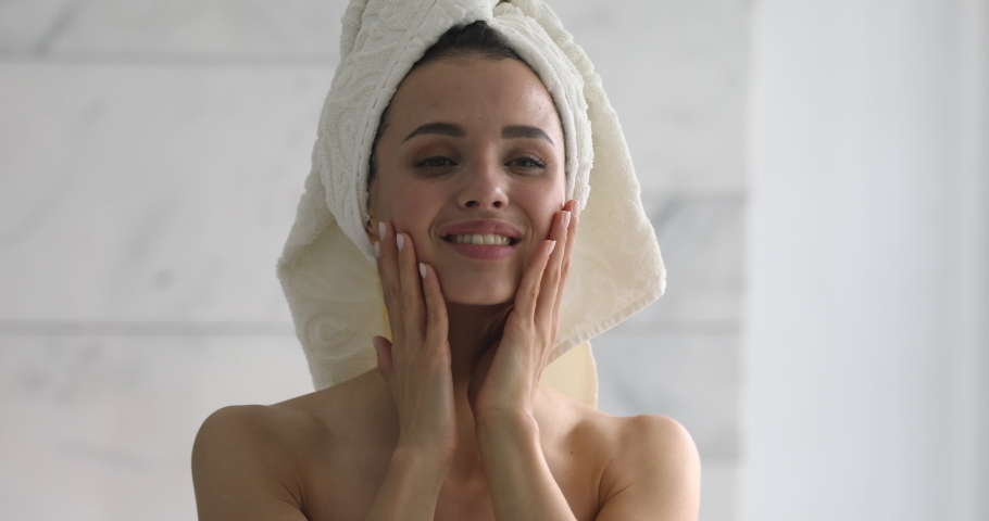 Happy attractive young woman with towel on head touch face after applying cream remove makeup look in mirror enjoy natural beauty and healthy clean soft moisturized skin care treatment, close up view Royalty-Free Stock Footage #1037145050