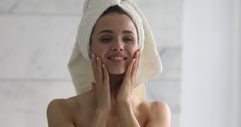 Happy attractive young woman with towel on head touch face after applying cream remove makeup look in mirror enjoy natural beauty and healthy clean soft moisturized skin care treatment, close up view