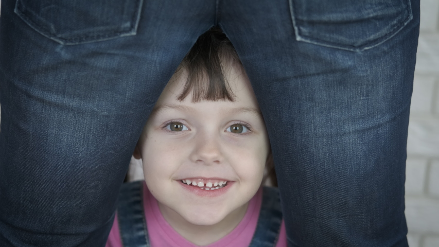 A cheerful child looks through the legs of a parent. Portrait of a happy little girl between mother