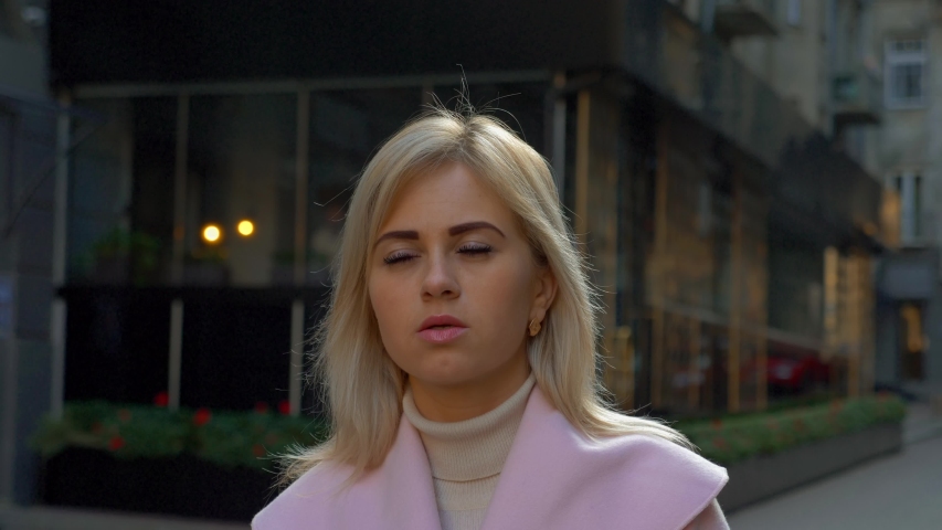 A young attractive blonde woman in a pink coat stands against the background of modern architecture. A beautiful girl is thinking while standing in the street. 