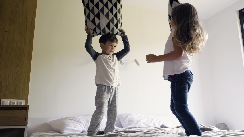 Adorable caucasian siblings have fun fighting with pillows on a bed in a sleeping room. Little boy and girl fooling around while parents away. Slow motion.