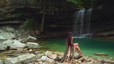 fantasy sexy siren mysterious brunette woman long hair green dress sits on white stones wild tropical lagoon waterfall. back no face Rear view. lake nature Georgia mountains Kutaisi turquoise water