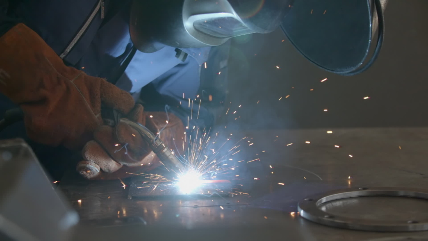 Close-up blacksmith welder in protective mask works with metal steel and iron using a welding machine, bright sparks and flashes in extreme slow motion