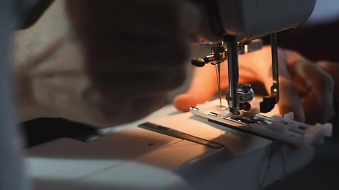 Focus on needle mechanism of sewing machine doing fast moves under light of built-in hardware lamp. Female proficient hands seamstress making decorative stitches on cloth detail using guide rails