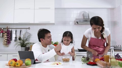 Happy Asian family make a cooking. Mother are preparing make salad, vegetable and Father with Daughter eating breakfast, cereal in the kitchen at home. Healthy food concept and happy holidays