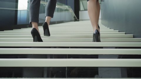Rear view closeup follow shot of two unrecognizable businesswomen in formalwear and high heels walking together upstairs in office building in slow motion