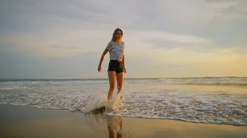 Young female felling fun and free and jumping barefoot on beach at sunset. Carefree girl on tropical beach runs into refreshing sea at golden sunrise. Happy tourist woman on summer vacation.