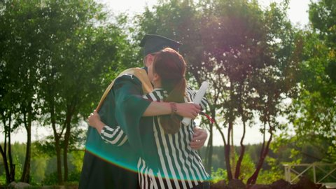 Excited Graduate Student in Gown and Cap with Diploma Hugs his Friend after Graduation Ceremony. 4K Slow Motion Static Medium Shot with Beautiful Sun Lens Flare