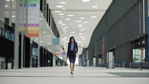 Lockdown wide shot of confident middle aged woman in suit and high heels shoes walking down office hall or airport lobby in slow motion 