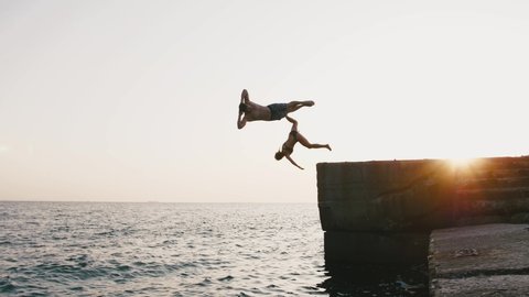Young woman and man jumping from a pier into the sea and doing tricks during beautiful sunrise, slow motion