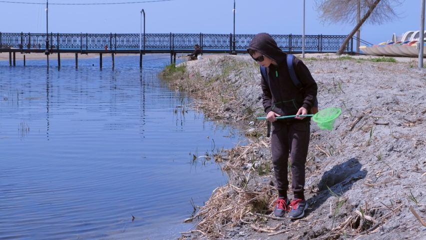 Boy is walking on the riverbank. He is catching frogs on the river using butterfly net but it jumps out. Young traveller with backpack. He is wearing sport suit and sunglasses. Child nature exploring.