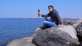 Man talks video chat at sea on smartphone with small tripod sits on stone. Young guy traveller shares his impressions about rest on mobile phone. Tourist at sea beach. Travel blogger work in journey.