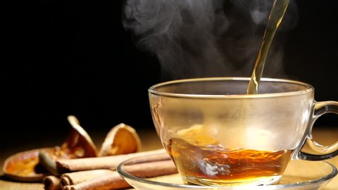 Pouring black tea or hot herb drink in a cup with steam in slow motion on black background