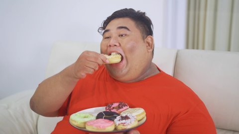 Overweight man enjoying a plate of sweet donuts while sitting on the sofa in the living room at home. Shot in 4k resolution