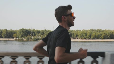Portrait of sportive athletic boy runs along city river. Side view of slim attractive man in activewear running fast on promenade. Muscular young guy running fast. Slow motion