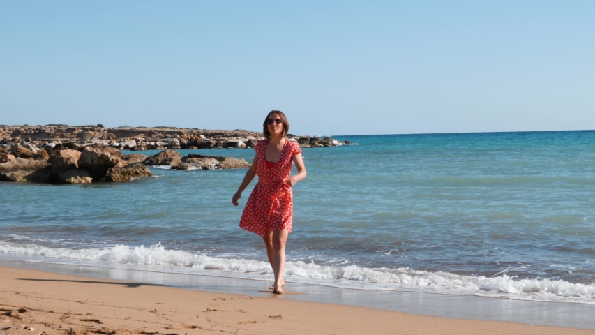 Young attractive woman walks along sandy beach. Brunette girl having fun on seashore. Girl basks in sun near sea. Girl relaxing on beach near sea with azure water. Girl has fun on vacation. Slow mo