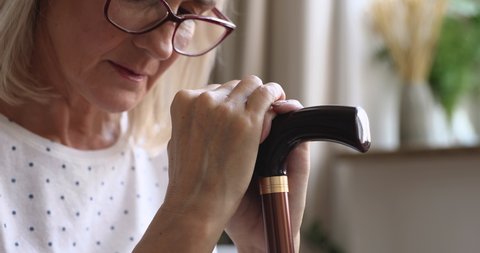 Upset old senior woman hold cane stick feel sad depressed about health problem sit alone at home near window wait caregiver visit, frustrated grandmother worried of disease concept, close up view
