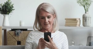 Happy middle aged mature woman holding smart phone looking at cellphone screen playing mobile games using social media apps in smartphone sit on couch at home, older people and technology concept