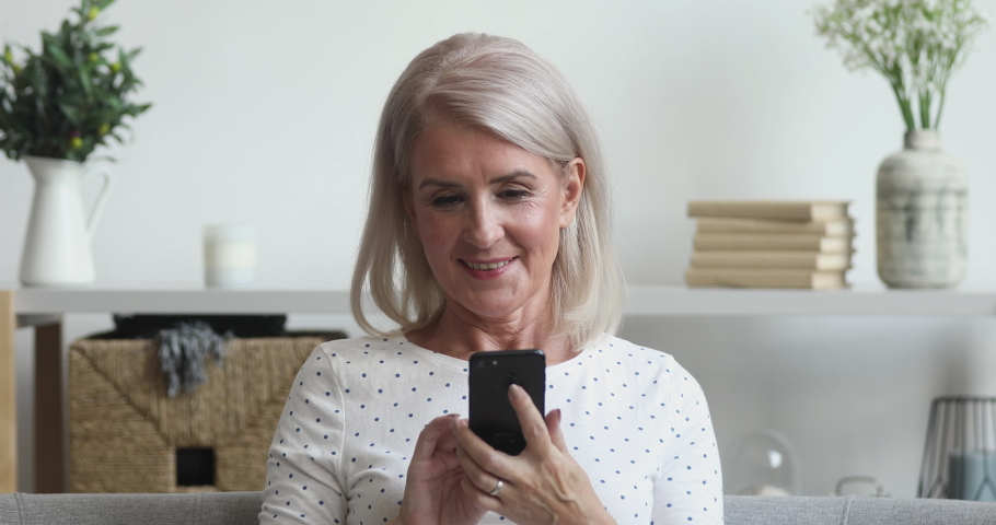 Happy middle aged mature woman holding smart phone looking at cellphone screen playing mobile games using social media apps in smartphone sit on couch at home, older people and technology concept | Shutterstock HD Video #1037252627