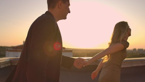 Young couple holding hands walking woman leading boyfriend the roof of the building at sunset POV travel concept. Carefree free lovers run on the roof laughing and smiling.