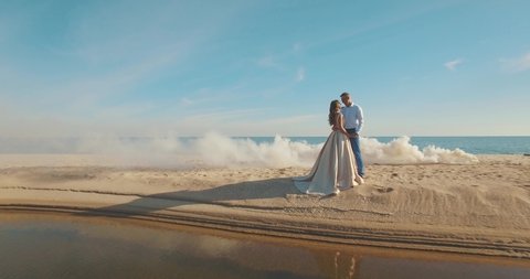 Aerial video of young wedding couple, on a clean sand beach shrouded by white fume. Viewing crystal clear blue sea from above, Drone flying out, 4K Footage