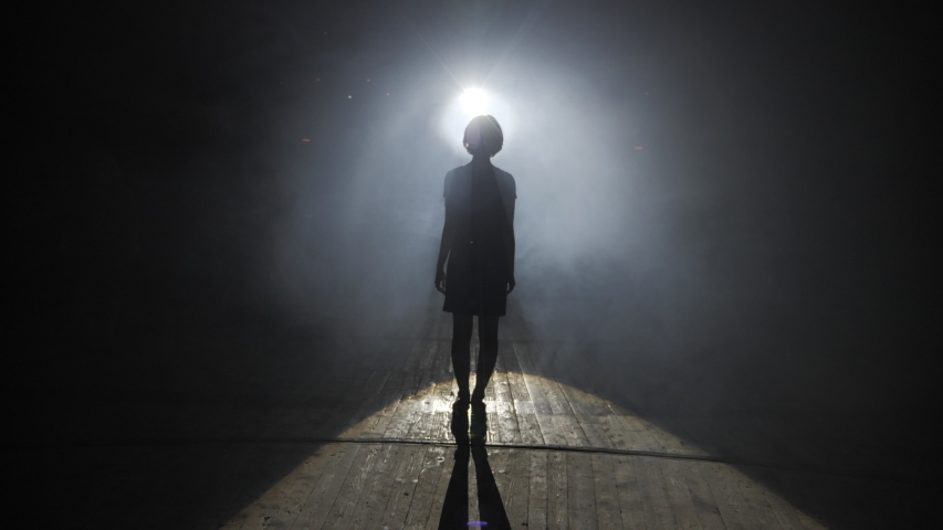 dark silhouette of young woman standing on wooden floor against bright spotlight creating halo in studio slow motion Royalty-Free Stock Footage #1037284358