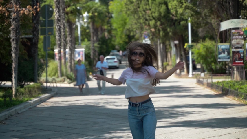 Young girl joyfully runs along the road. A happy young girl in sunglasses runs along the road, waves her hands and smiles, slow motion.
