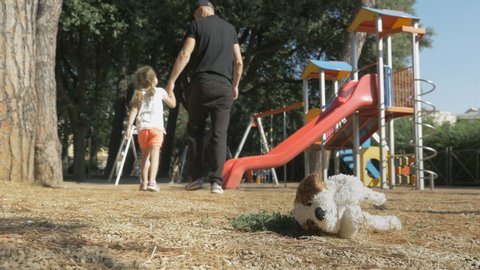 A man in black clothes leads a child out of the playground. Children's soft toy lies on the ground in foreground. Stranger abducted a child