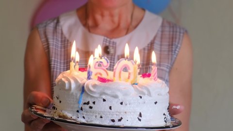 Mature woman's birthday. Cheerful elderly woman blows out the candles on the cake, close up, slow motion