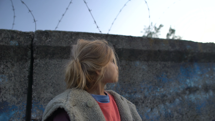 Little girl with muddy face standing at barbed wire, war child in refugee camp