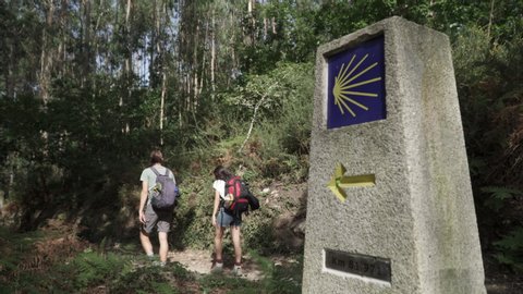 Way of St. James sign and pilgrims walking on a forest footpath. Yellow scallop sign pilgrimage to Santiago de Compostela