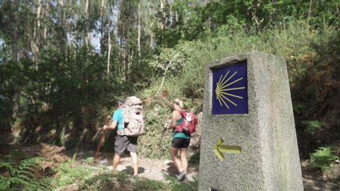 Camino de Santiago sign and two pilgrims walking on a forest footpath. Yellow scallop sign pilgrimage to Santiago de Compostela